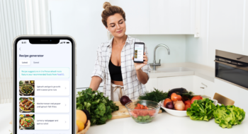 Woman surrounded by fresh product in the kitchen holds up mobile phone with Recipe Generator